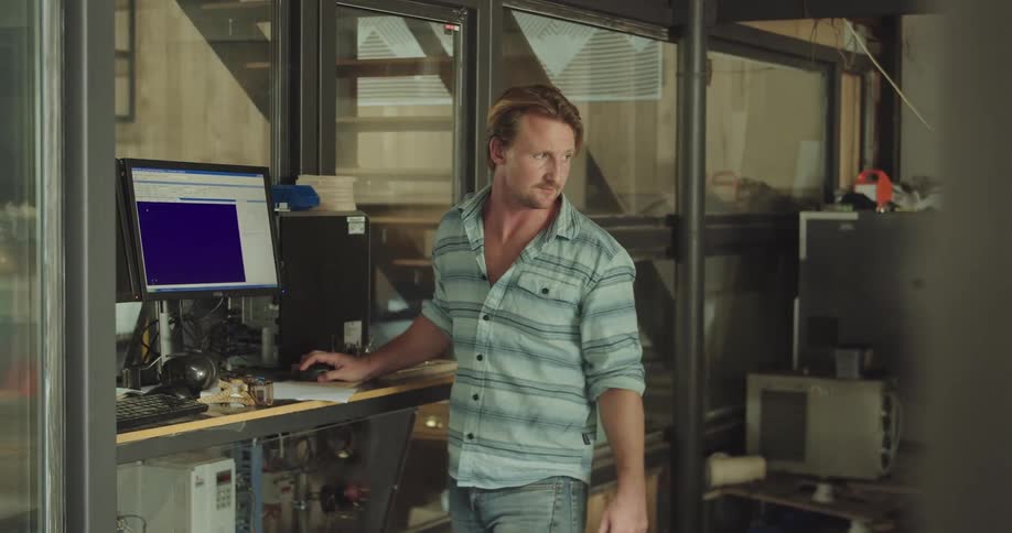 A man stands at a computer and turns to tell someone something in a workshop