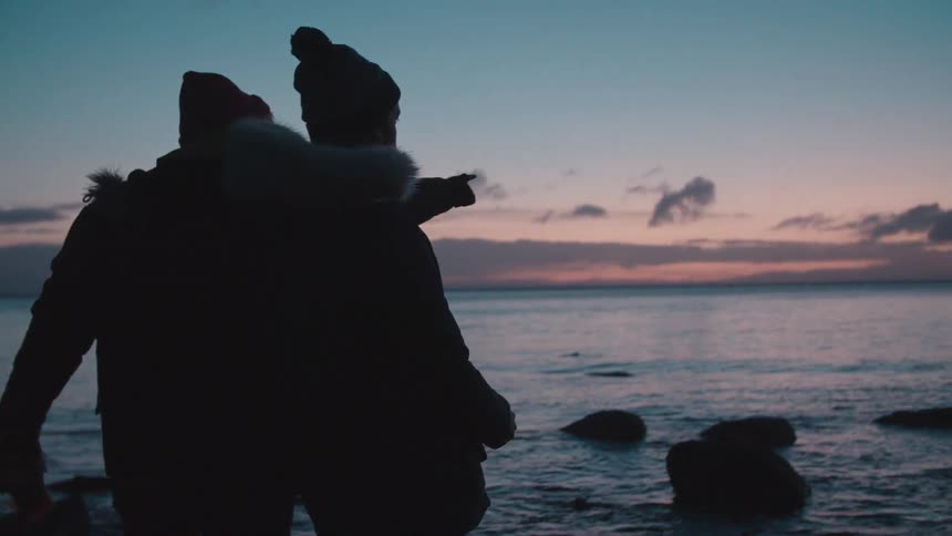 A person walks over some rocks by the shore to point something out to a friend at sunset