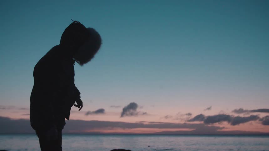 A person wearing a parka throws a stone into the ocean at dusk