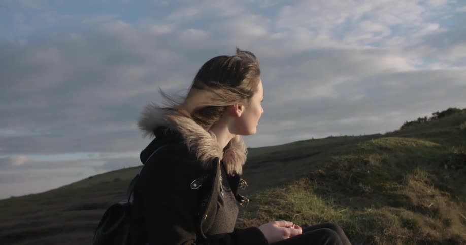 A woman sits on a windy hillock thinking and looking outward