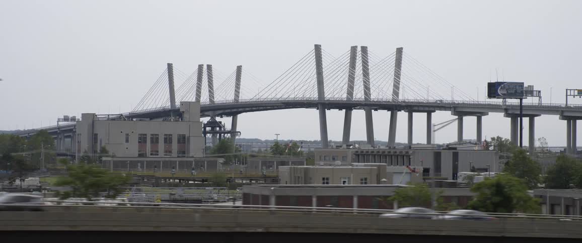 Cars drive down highway on a cloudy day with a bridge trees and buildings in the back ground