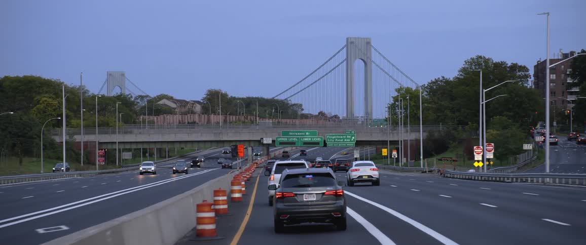 Cars drive down highway in the evening past trees and buildings toward a bridge and under an over pass