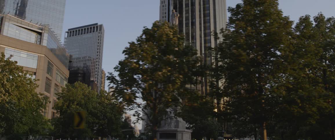 A stone monument with a statue at the base is in the city center surrounded by trees