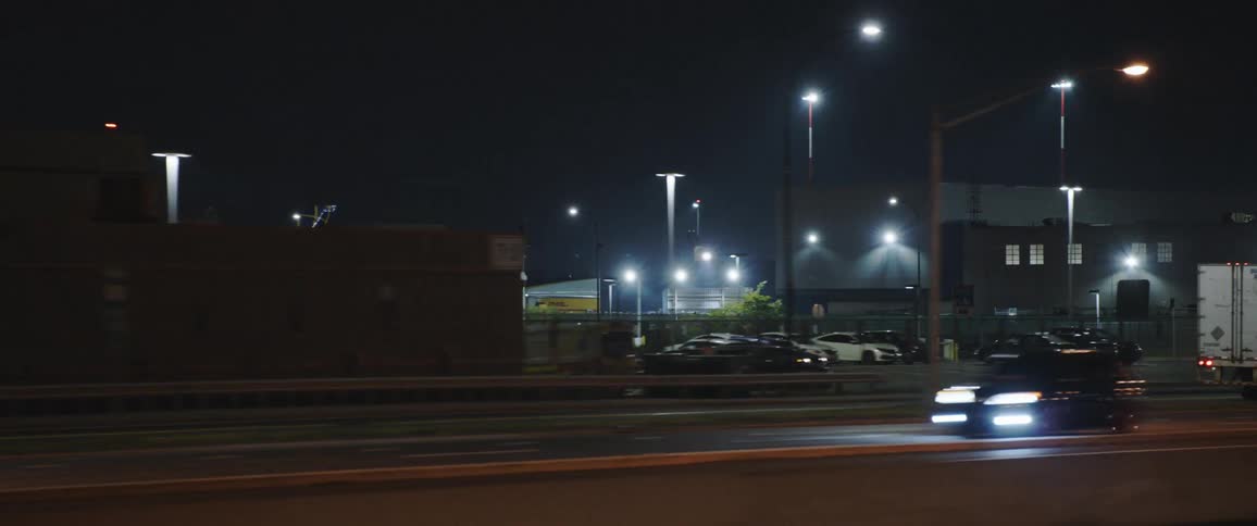 Cars drive down high way at night with buildings and parked cars in the background