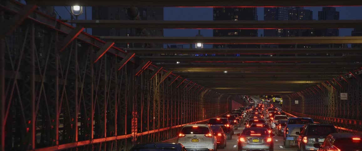 Cars drive down a road under a metal structure toward a city at night