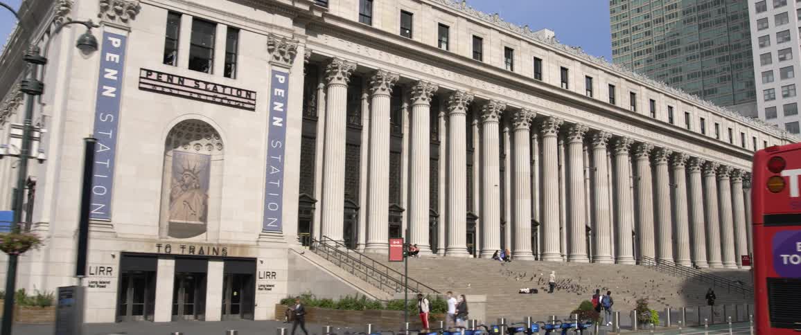 People walk past and hang out on the steps of penn station while other people ride by on a tour bus with buildings in the back ground on a sunny day