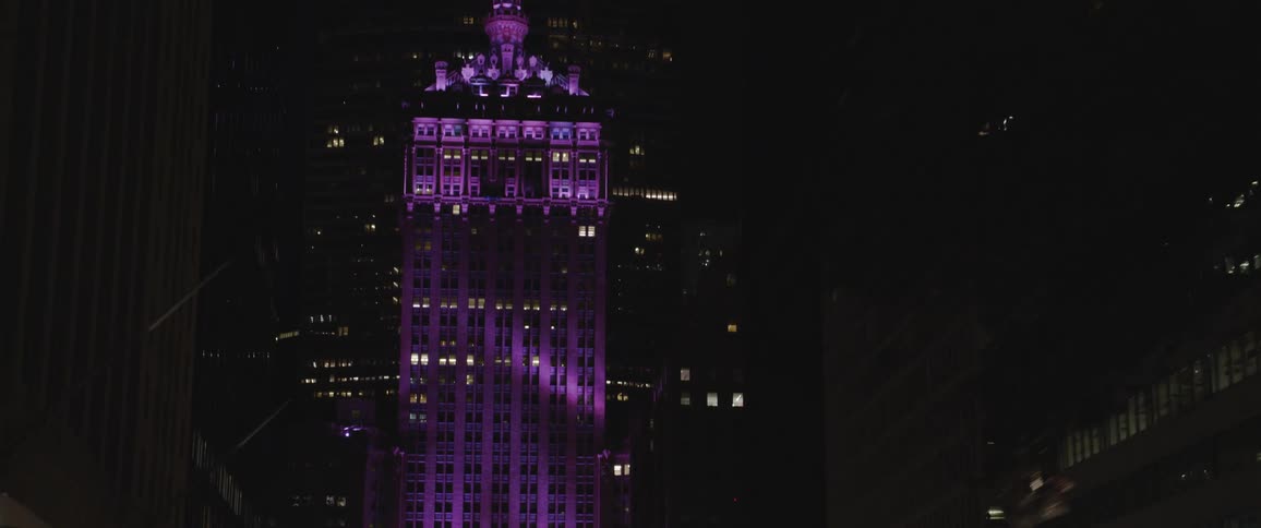 Street vantage of downtown with buildings trees street lights at night with a building illuminated in purple light