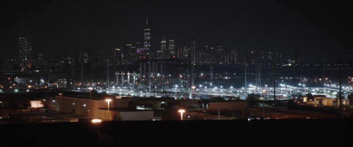 Cars drive by a factory with the city skyline behind it