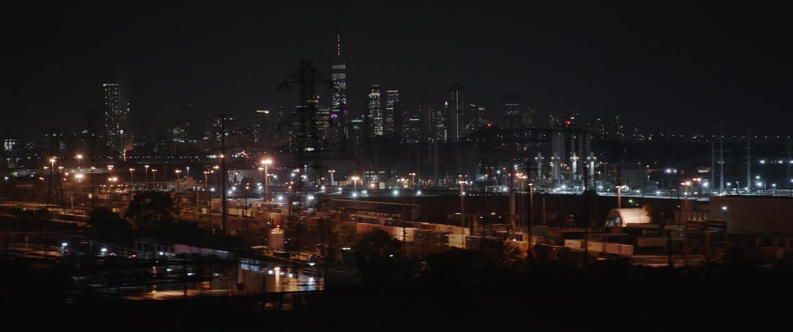 Vehicles driving past a factory with a city skyline in the distance