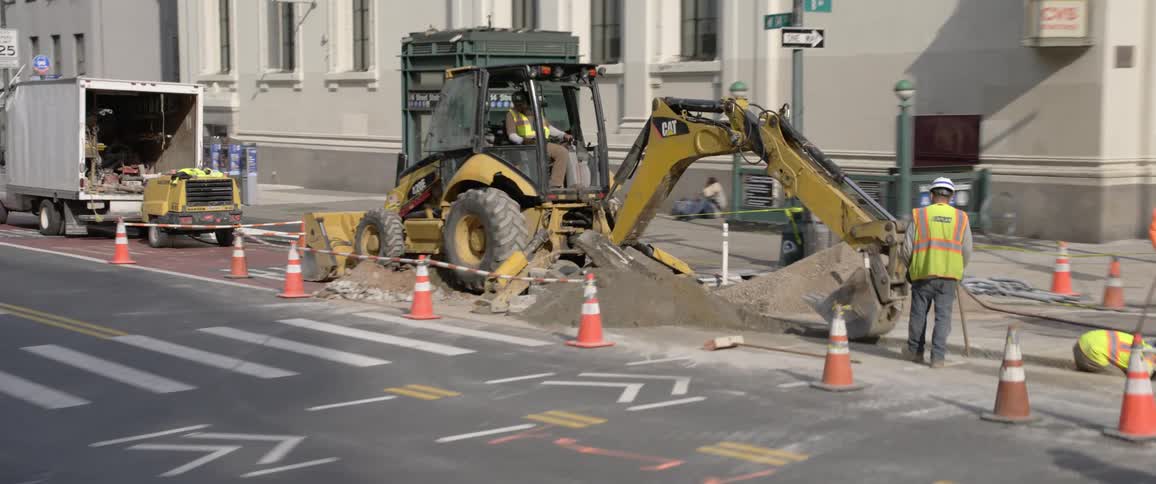 Men dig a hole and work next to a subway entrance an intersection and a building in a downtown area on a sunny day