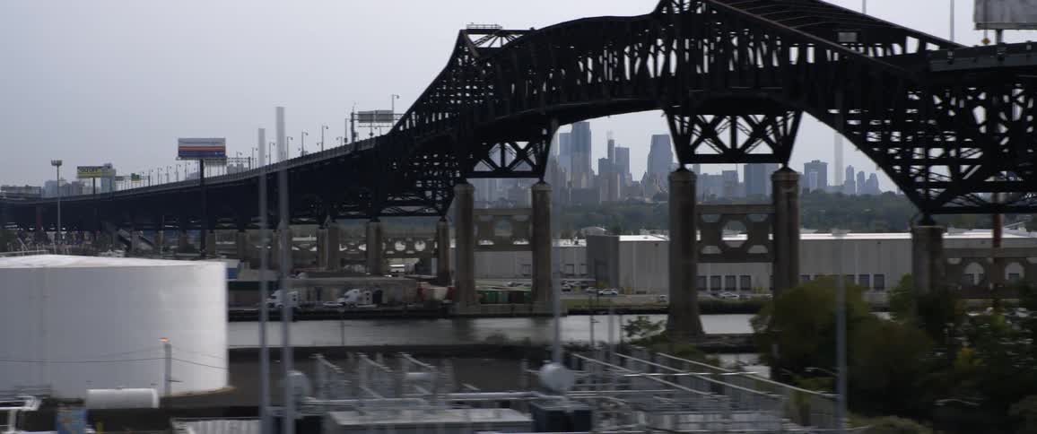Street parallels river passing bridge and refinery across from skyline on misty day
