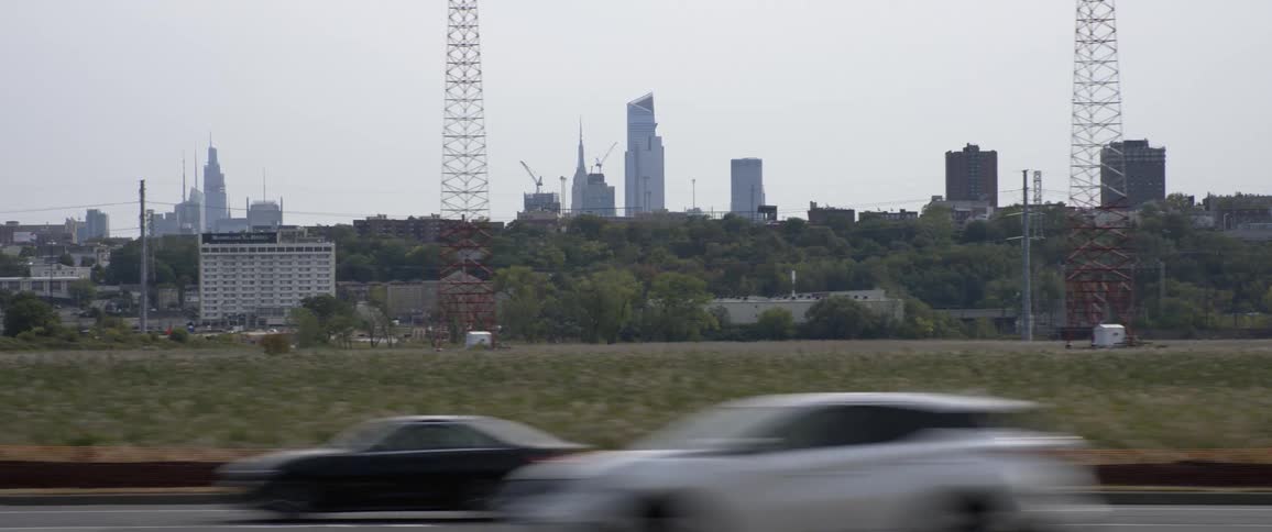 Cars drive down road past towers trees and grassy areas in front of a city skyline on a sunny day