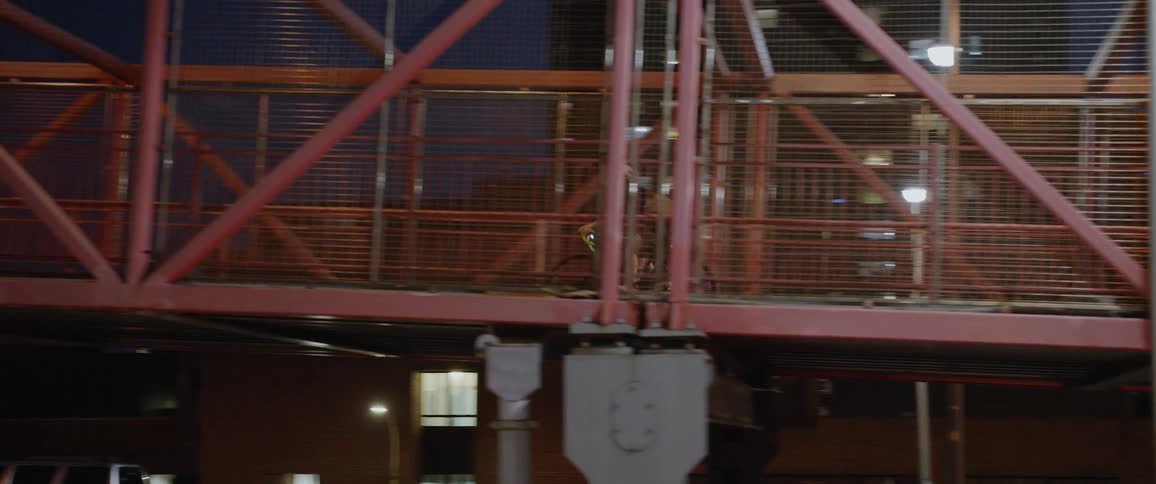 People ride bikes or walk on a bridge in a cage above cars driving on a road at night