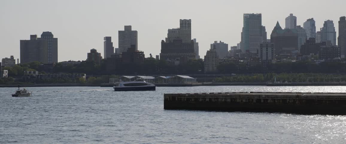 Boats go across water behind pier and in front of city skyline
