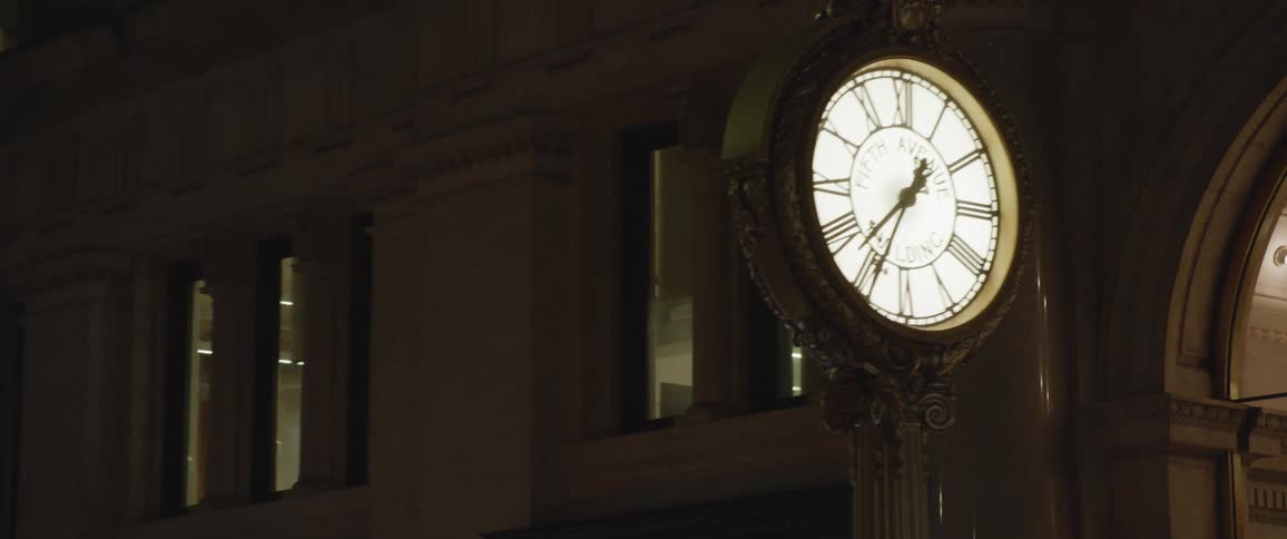 A clock with an internal light outside of a building with an arch and windows