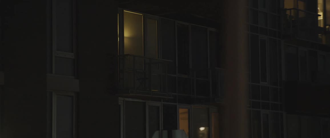 Apartment building at night with lights on in some apartments and a tv screen in one all have a balcony