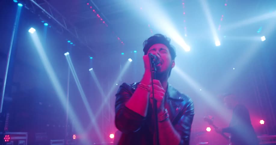 A man stands on stage singing into a microphone with two band members playing instruments behind him on a stage with spotlights
