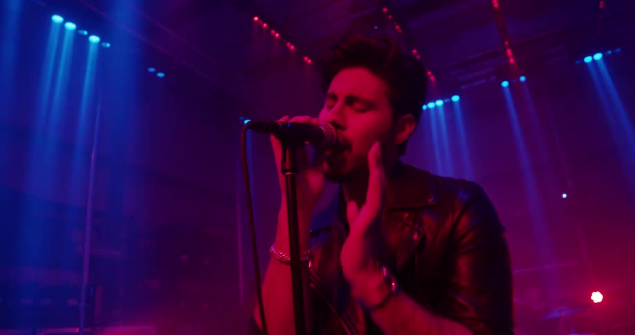 A man singing on stage with his band playing instruments behind him