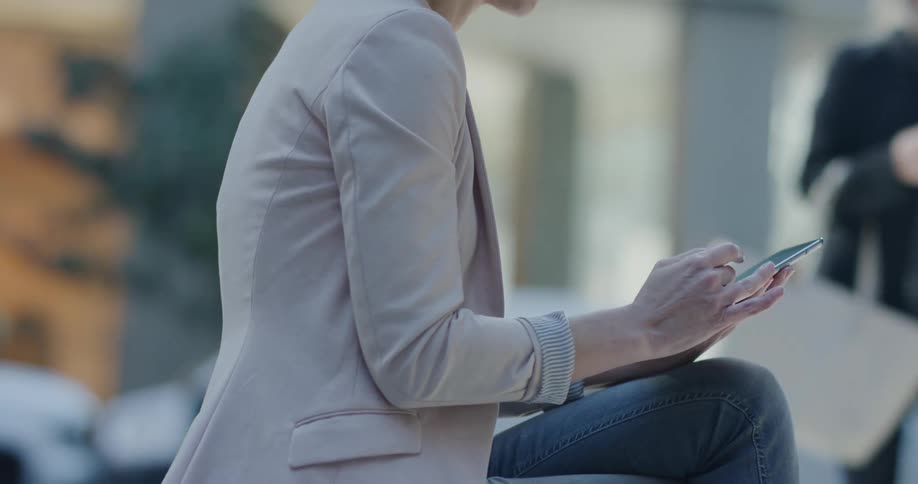 A woman sits and uses her smartphone on a bench downtown as people walk past