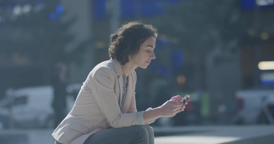 A woman sits outside on a busy city street texting on her cell phone