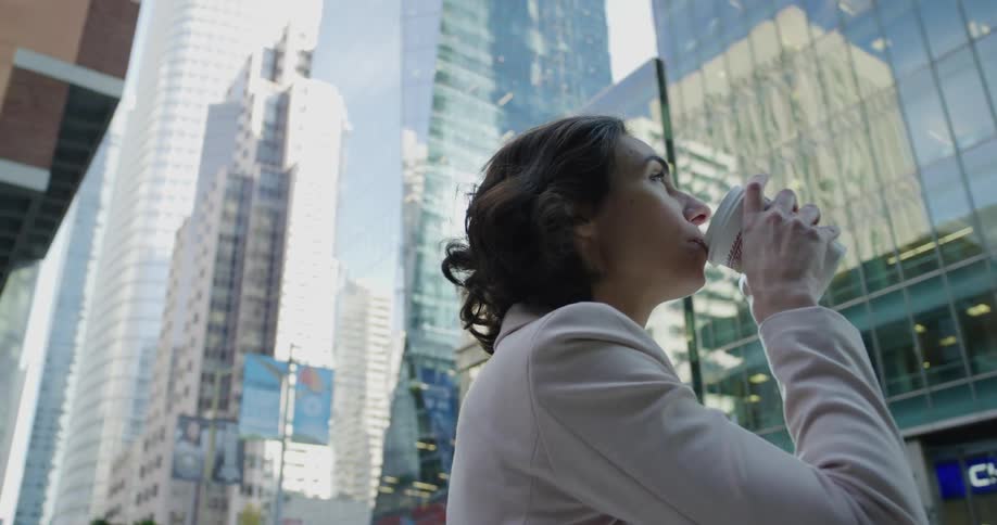 Woman with pink blazer jacket drinks coffee in downtown with high rise buildings