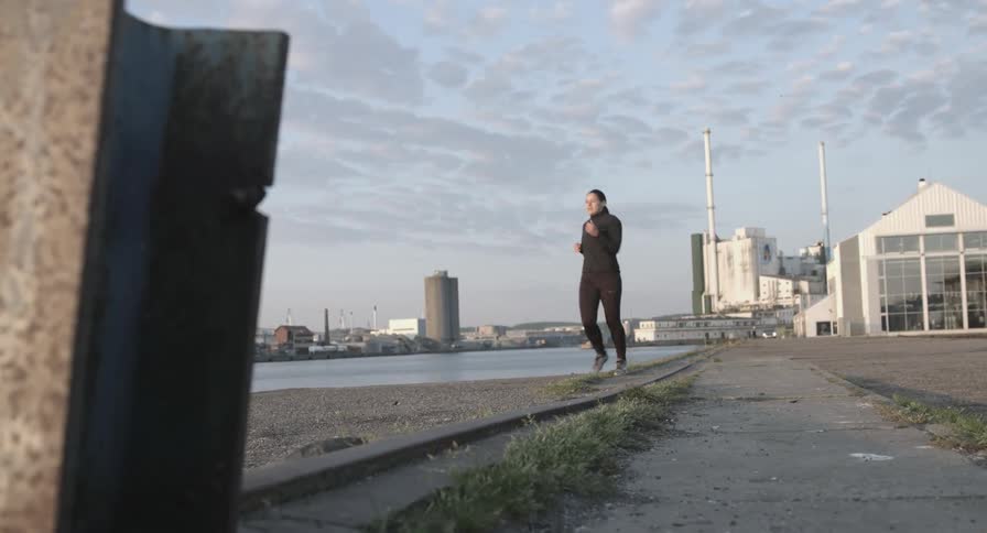A woman jogs to a stop on a path near a river in an industrial era