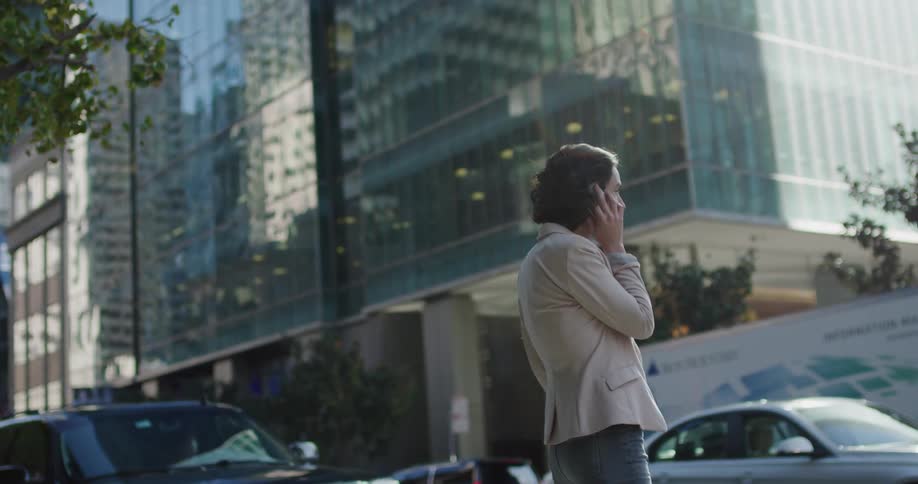 A young woman talks on her smartphone while standing outdoors beside a road in a metropolis during the day