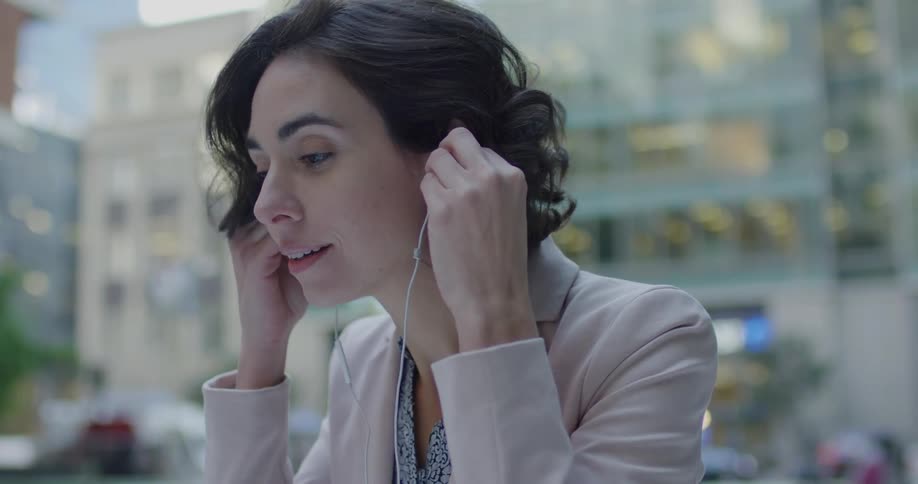 Woman sitting outdoors in a downtown area puts a pair of white earbuds in her ears and smiles