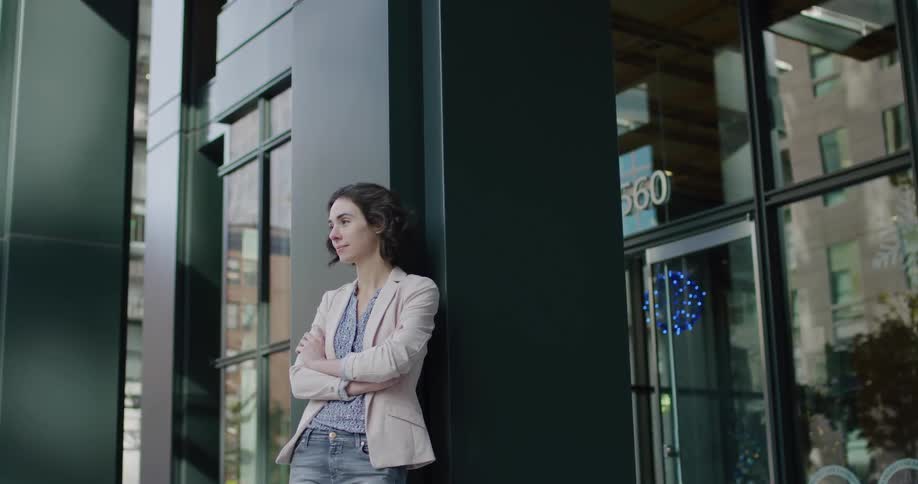 A woman stands outdoors with her back against the pillar of a building in a city during the day
