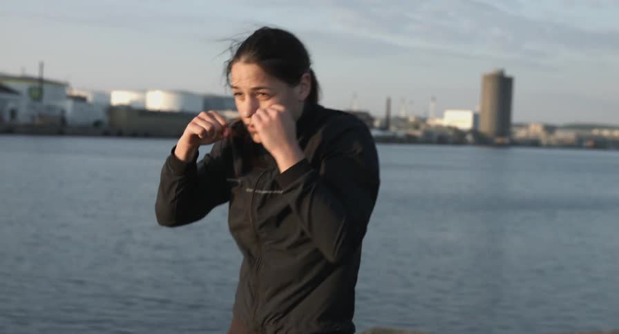 A woman practices her punching maneuvers outside in front of a body of water and buildings