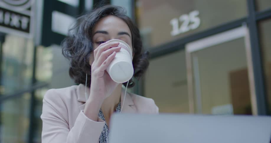 Brunette woman with pink blazer jacket and earphones drinks coffee while using laptop outside of coffee shop