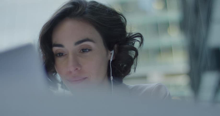 A woman sits inside working on a laptop while wearing earbuds