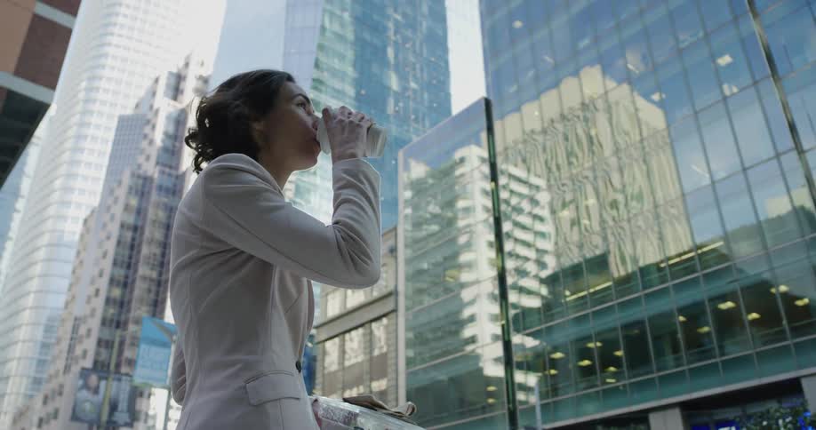 A woman stands outside an office building and drinks a coffee while holding a salad
