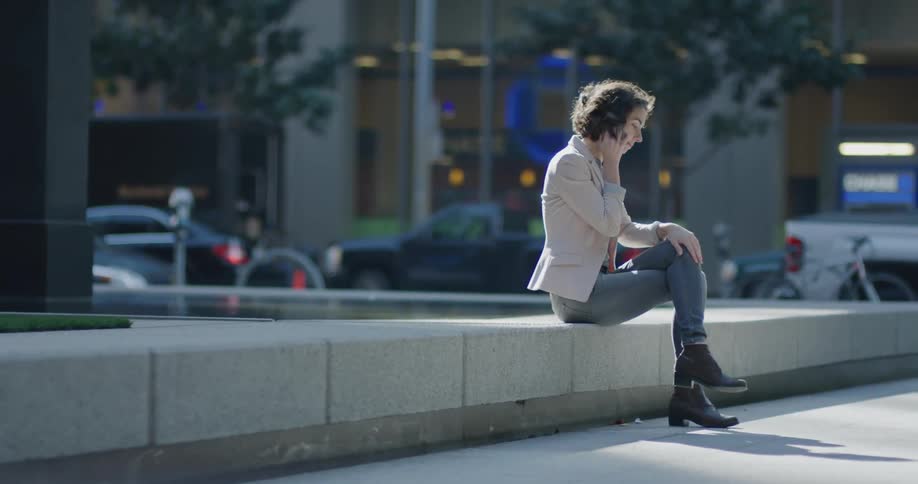 Woman talks on a cellphone while sitting on a cement block as traffic and pedestrians move behind her