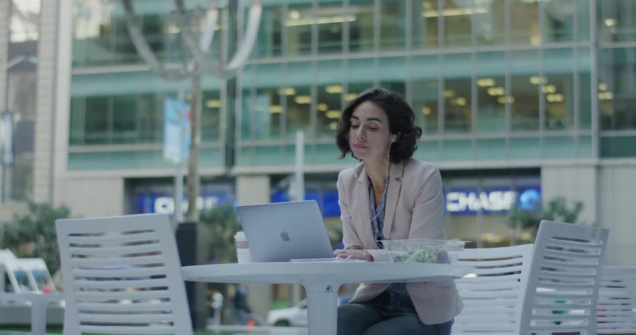 Woman sits at a white table outside in an urban area eating a salad while working on an apple laptop computer