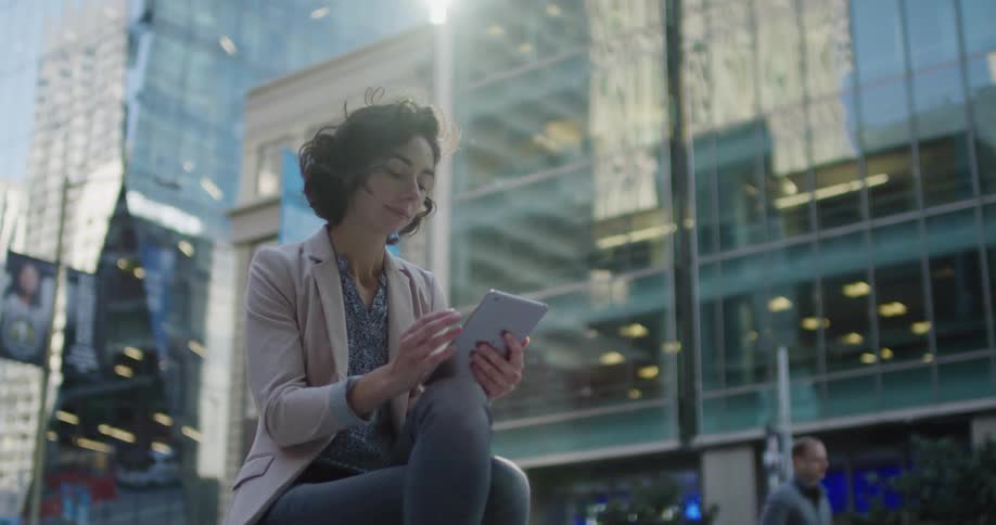 A woman scrolling on a tablet while sitting near large city buildings on a sunny day