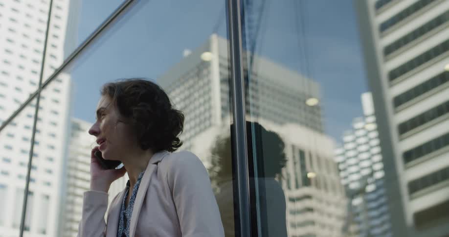 A woman standing with her back against a window on the facade of a building speaks on her smartphone on a sunny day