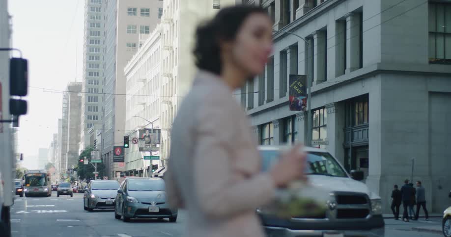 A business woman waiting to cross the street in a downtown city while holding onto her coffee and salad