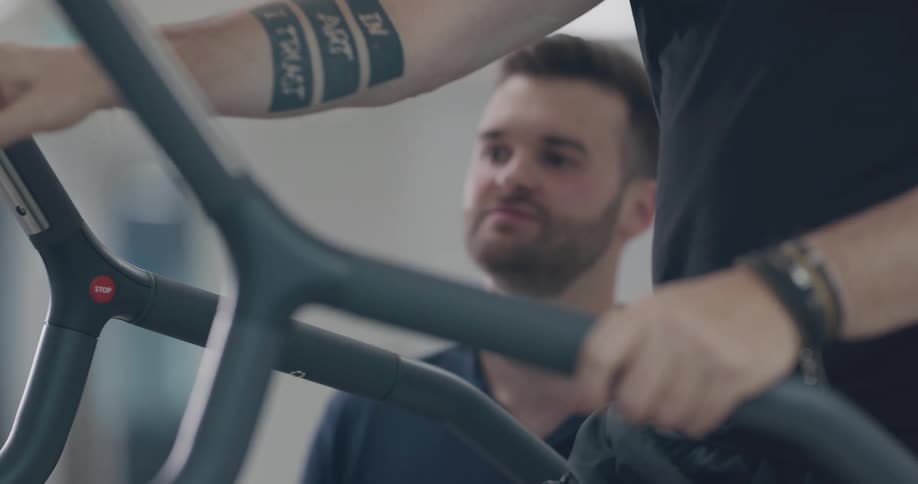A man wearing black steps onto exercise equipment as a male trainer looks over him in a gym