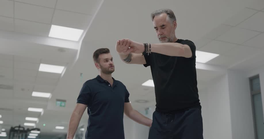 A fitness trainer helps an older man perform a stability exercise within a gym