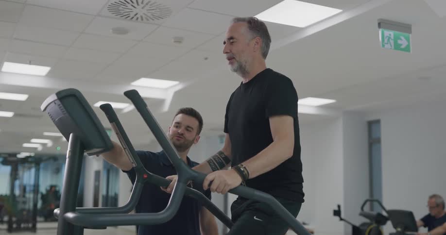 A man with a beard and wearing a black outfit exercises on a piece of equipment with a trainer standing beside him