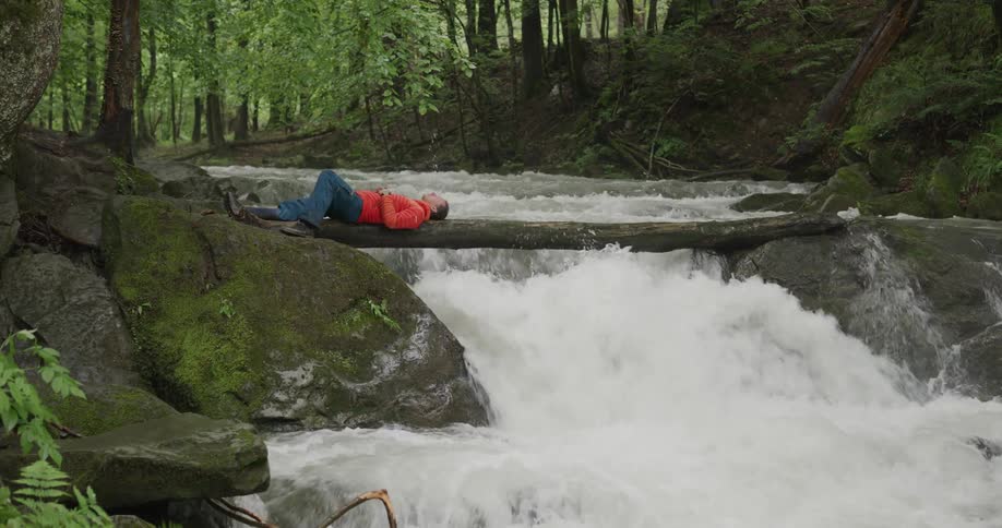 Man in red jacket laying on a log over a river in the forest on the mountains