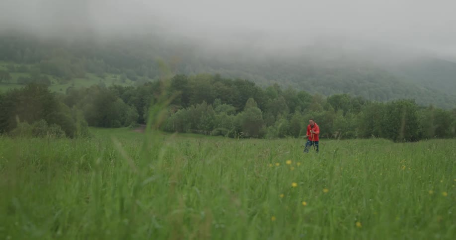 Man wearing orange jacket walking out of forest through the grass on a foggy day