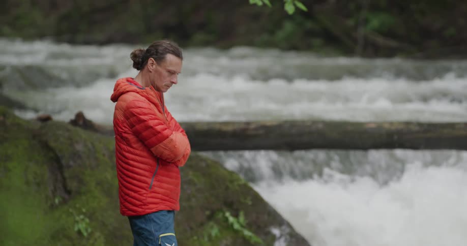 A man in an orange coat standing next to a gushing river in nature