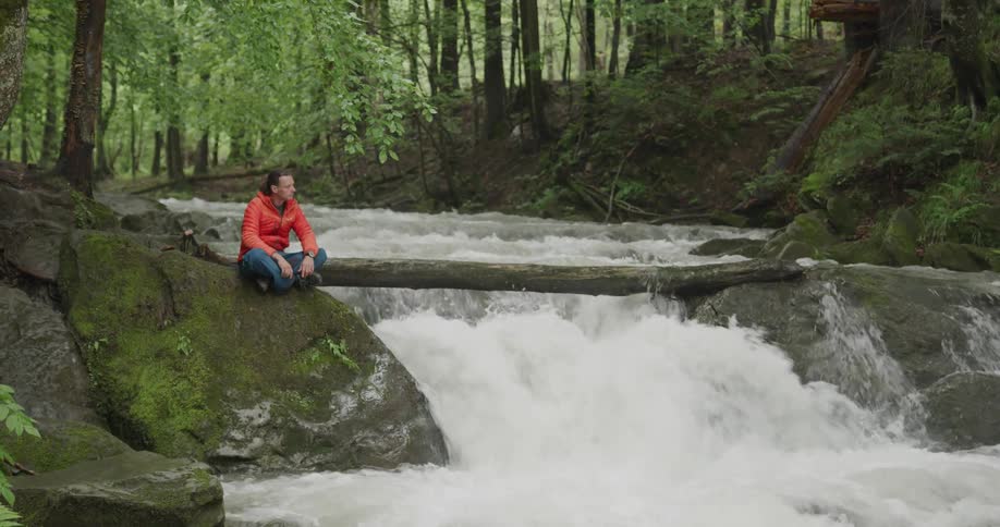 Man sits cross legged on a rock next to the water splashing over the rocks in a river