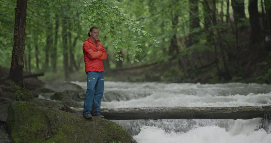 A man with a jacket standing next to a river with trees in the background