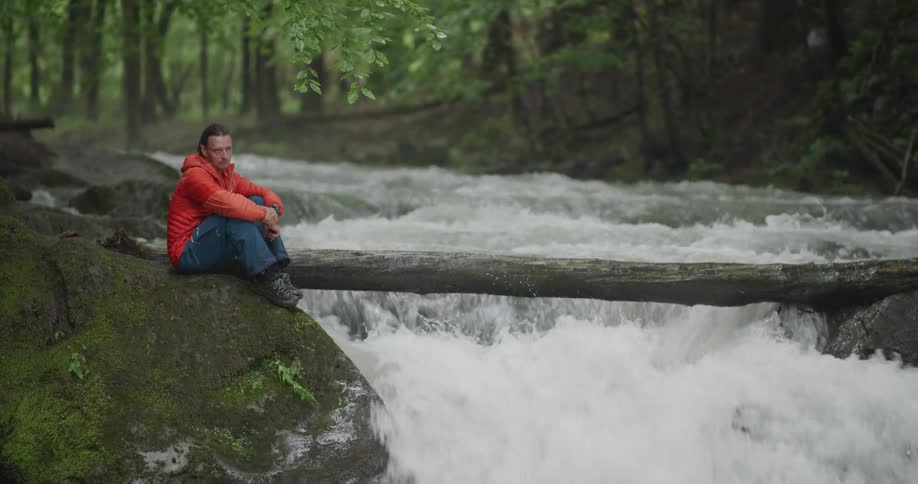 A man sits at the edge of a rushing river that is flowing swiftly through a forest