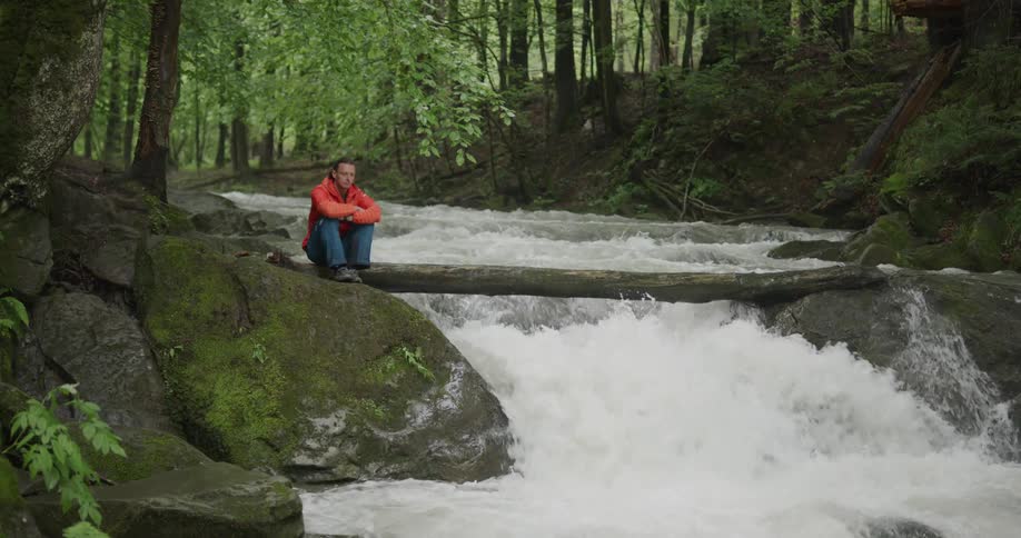 Man sitting on a log at a stream watching water rapidly flowing down