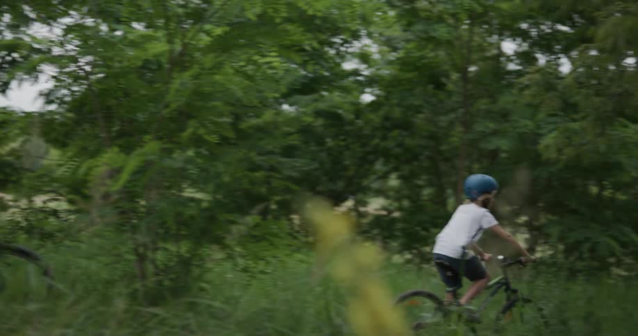 A boy and a man ride together on bicycles through a woodland during the day