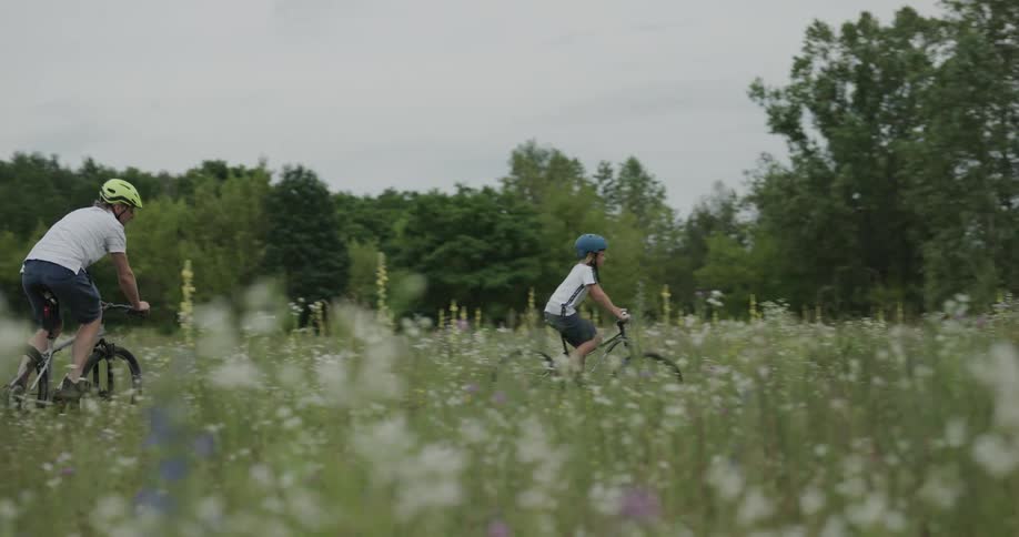 A boy and a man ride their bicycles through a field of tall grass on a cloudy day